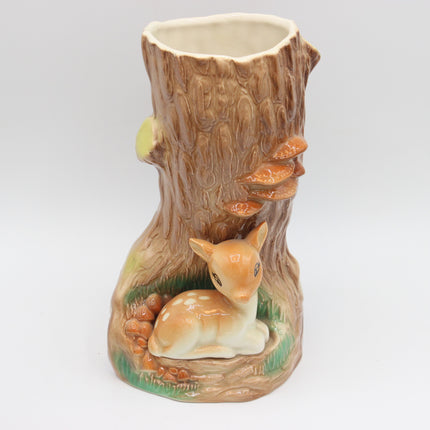 A ceramic tree stump planter featuring a small white and tan deer figurine nestled at the base, with brown mushrooms and green grass details, set against a plain light gray background.