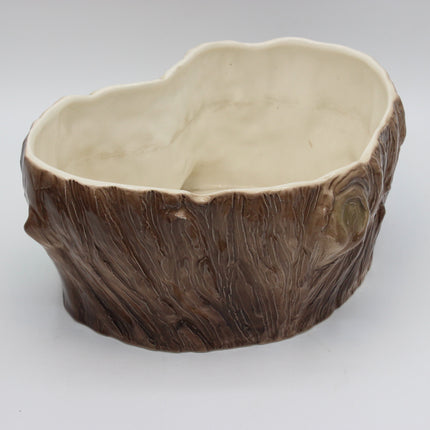 A ceramic planter shaped like a tree trunk, featuring a textured brown bark exterior and a smooth, cream-colored interior, shown on a neutral light gray background.