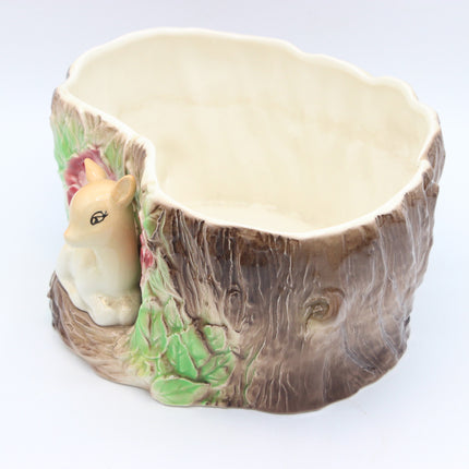 A ceramic woodland-themed planter shaped like a tree stump, featuring a cream-colored interior, brown bark-textured exterior, green leaves, and a small beige animal figurine on the side.