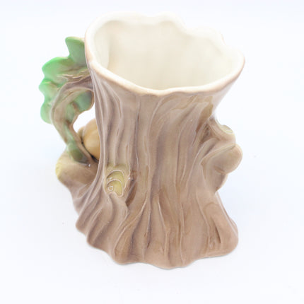 A ceramic planter shaped like a tree trunk, featuring a textured brown bark design, a green leafy branch on the side, and a light beige interior, set against a plain white background.