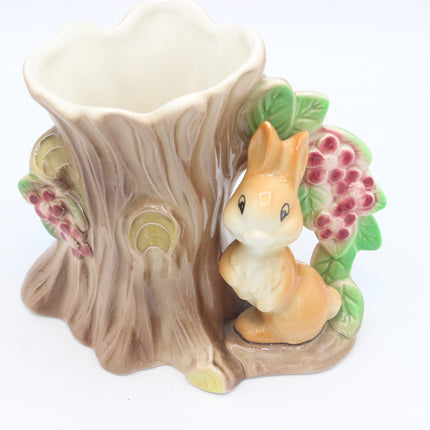 A ceramic bunny and tree stump holder with a small orange rabbit figurine beside a tree trunk-shaped cup, decorated with green leaves and pink berry clusters on a white background.