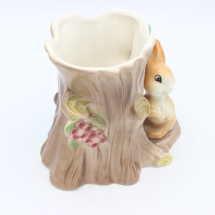 A ceramic tree stump planter with a small painted bunny figure on the side and floral details, shown on a plain white background.