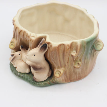 A ceramic planter shaped like a tree stump with three cream-colored rabbits nestled underneath, set against a plain white background.