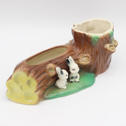A ceramic tree stump planter with two small white rabbits sitting on a green leafy base, photographed on a clean white background.