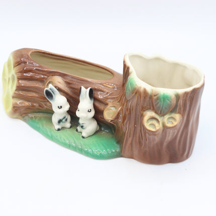 A ceramic woodland-themed plant pot shaped like a tree stump and log, featuring two small white rabbits on a green base, photographed on a white background.