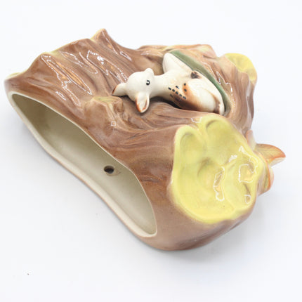 A ceramic decorative pot shaped like a tree trunk, featuring a glossy brown textured surface, a yellow glazed patch, and a small white animal figurine nestled in the center, shown on a plain white background.
