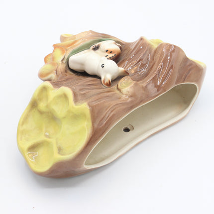 A ceramic dog poop bag shaped like a log, featuring a yellow-green paw print base, brown textured bark, and a small white dog figurine peeking out from the top, shown on a white background.