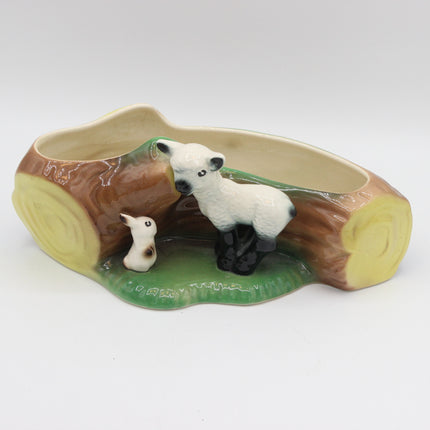 A ceramic planter shaped like a log with a white lamb and a small duck figurine on a green grassy base, photographed on a light gray studio background.