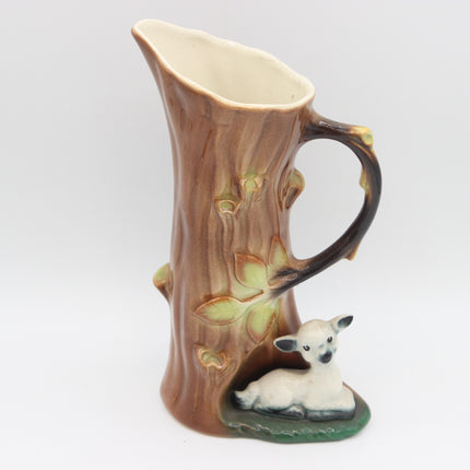 A ceramic pitcher shaped like a tree trunk with a curved handle, featuring a small white lamb figurine sitting at the base on a green surface, photographed against a plain light gray background.