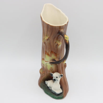 A ceramic mug shaped like a tree trunk with a handle, featuring a small white lamb figurine at the base, photographed against a neutral light gray background.