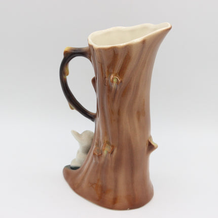 A ceramic pitcher shaped like a tree trunk, featuring a brown and tan glazed finish with a dark brown handle and a small white animal figure at the base, shown on a light gray background.