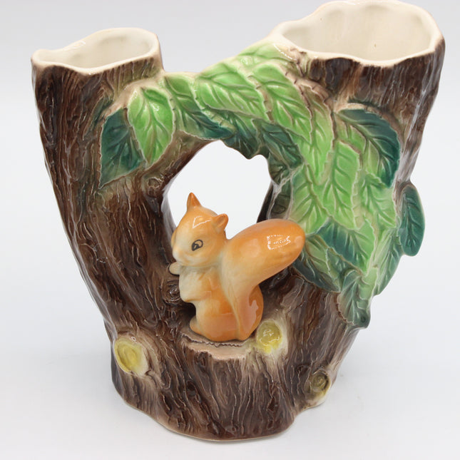 A ceramic double cup holder designed to look like a tree stump with a small orange-brown squirrel figurine in the center, flanked by green leaves and two open cup slots at the top, on a white background.