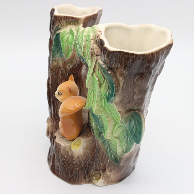 A ceramic two-bottle vase designed to look like a tree stump, featuring a small glazed squirrel figure and green leaves on the front, set against a plain white background.