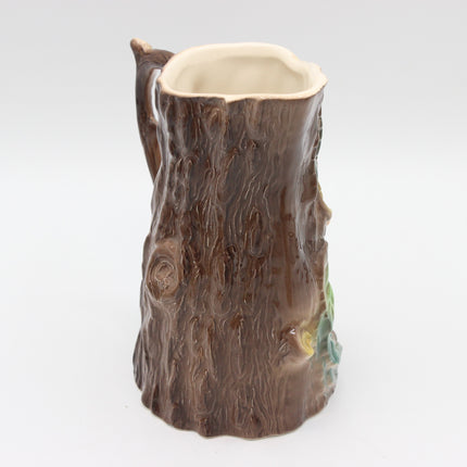 A ceramic pitcher shaped like a tree trunk, featuring a textured brown bark design, a small side handle, and a light cream-colored interior, set against a plain white background.