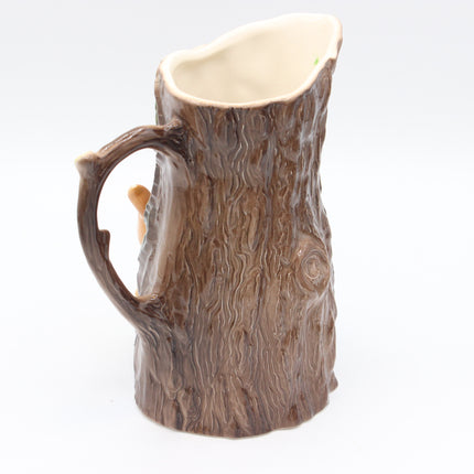 A ceramic pitcher shaped like a tree trunk, featuring a textured bark design in brown tones and a cream-colored interior, set against a plain white background.
