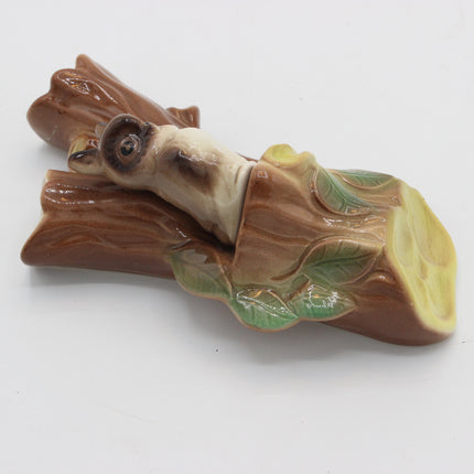 A ceramic decorative figurine of a brown tree stump with a small owl perched on top, featuring green leaves and a yellowed cut end, photographed on a white background.