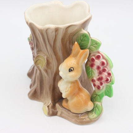 A ceramic tree stump flower pot featuring a small orange-brown bunny figurine, green leaves, and pinkish-red berries on a white background.