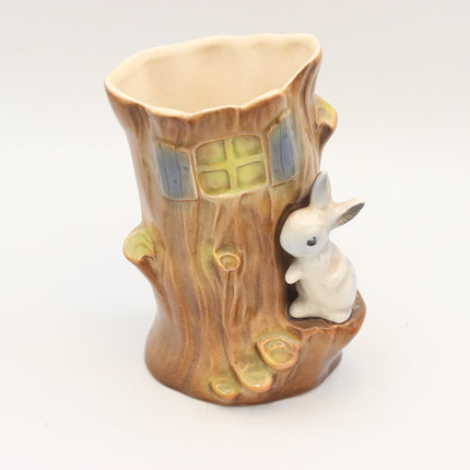 A ceramic tree stump cup featuring a small white rabbit figurine on the side and a window detail on the front, set against a plain white background.