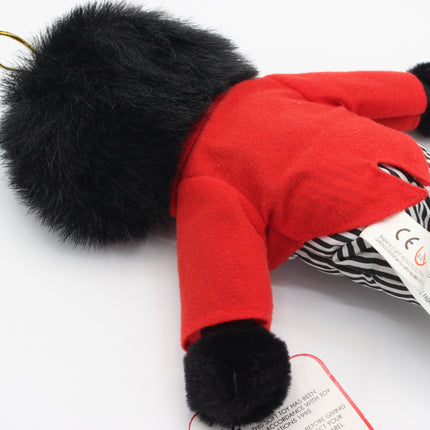 A close-up of a plush toy wearing a bright red jacket and black-and-white striped trousers, with a large black faux-fur pom-pom on one side and a white product tag attached.
