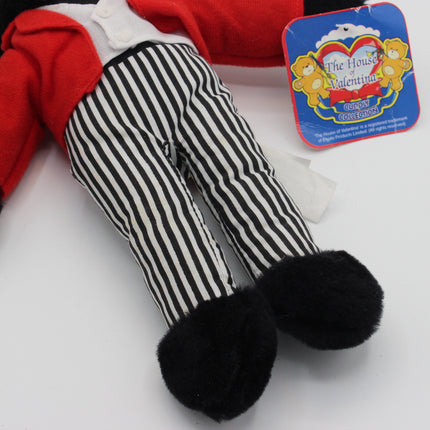 A plush doll or toy Golly figure shown from the waist down, wearing black-and-white vertically striped pants, a red jacket, and black fuzzy shoes. A blue hangtag reading 'The House of Valentine' is attached near the upper left.