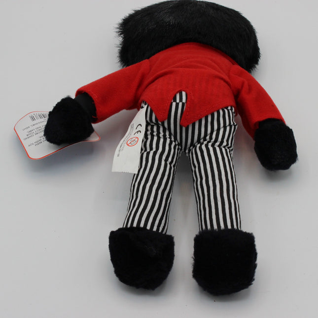 A small plush doll shown from the back, wearing a bright red top, black-and-white vertically striped pants, and black fuzzy cuffs and feet, photographed on a plain light background with a paper tag attached.
