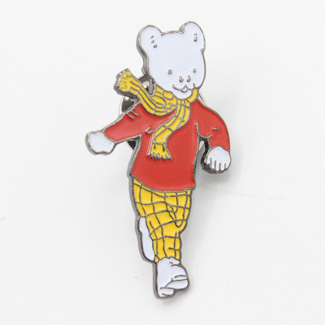 A small enamel pin shaped like a walking rupert bear wearing a red top, yellow plaid pants, and a yellow scarf, shown on a plain white background.