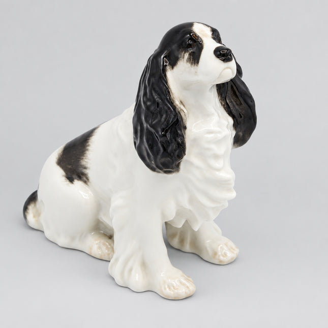 A ceramic figurine of a black and white English Cocker Spaniel dog sitting down, photographed against a seamless white background.