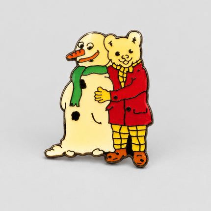A small enamel pin depicting a yellow bear hugging a snowman, shown on a plain white background.