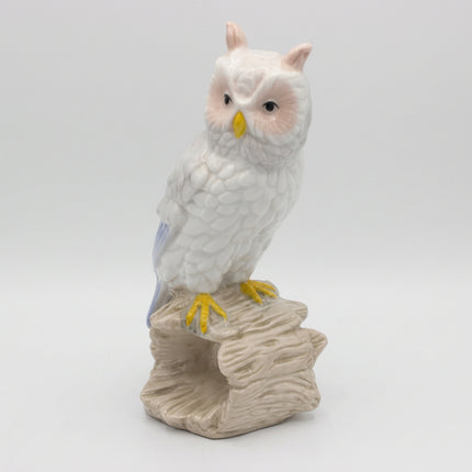 A white owl figurine with yellow beak and feet, perched on a textured beige branch or log base against a light gray studio background.