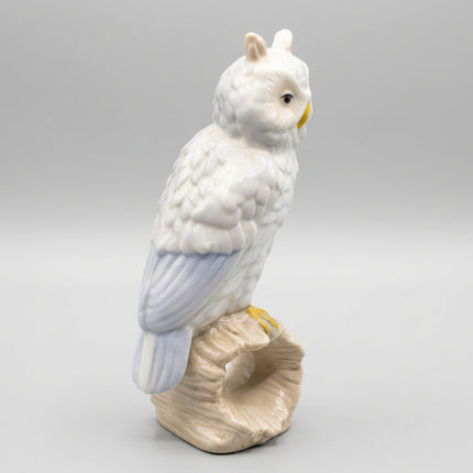 A ceramic owl figurine perched on a branch, featuring textured white feathers, soft blue wing accents, and a yellow beak and feet, set against a plain light gray background.