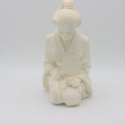 An off-white alabaster  figurine of a kneeling woman in traditional robes holding a round fan, set against a seamless light gray background.