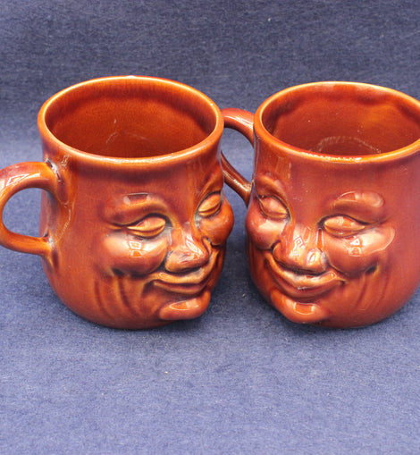 Two brown ceramic mugs with face designs on a blue background