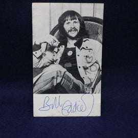 Black and white photo of Bill Oddie with long hair and a jacket, sitting on a chair.
