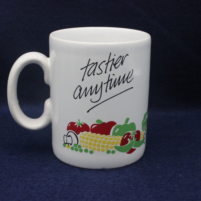 White mug with 'tastier anytime' text and vegetable graphics on a dark background