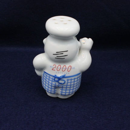 White ceramic figure with a chef's hat and blue apron, holding a saltshaker, on a dark background.