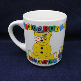 Pudsey Bear mug with a cartoon bear design and colorful text on a dark blue background
