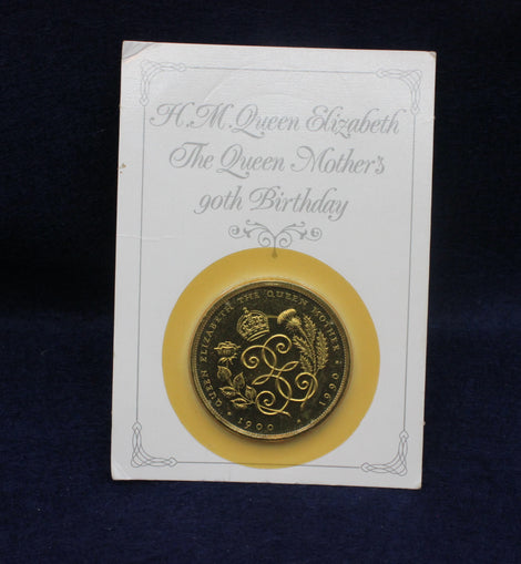 Gold coin on a card with text about Queen Elizabeth's Mothers 90th birthday, against a dark background.