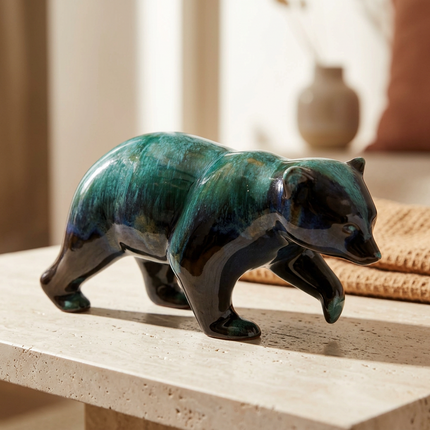 A glossy ceramic bear figurine in deep teal and black glaze, standing on a light stone tabletop in a softly lit interior setting.