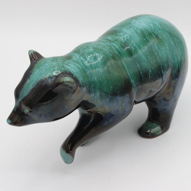 A glossy ceramic figurine of a bear in a walking pose, featuring a textured teal and dark blue-green glaze with black accents on the paws and face, set against a plain light gray background.