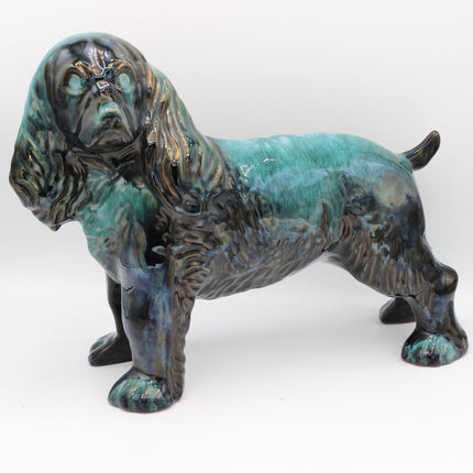 A glazed ceramic figurine of a long-eared dog, likely a Cocker Spaniel, featuring a textured dark brown and black body with a glossy turquoise and teal glaze, set against a plain white background.