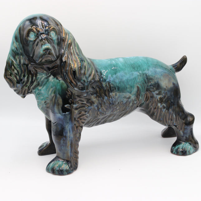 A glazed ceramic figurine of a long-eared dog, likely a Cocker Spaniel, featuring a textured dark brown and black body with a glossy turquoise and teal glaze, set against a plain white background.