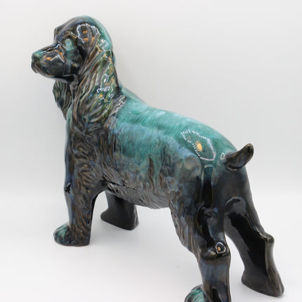 A ceramic figurine of a spaniel dog with a glossy teal, green, and black glaze, standing on a light gray background.