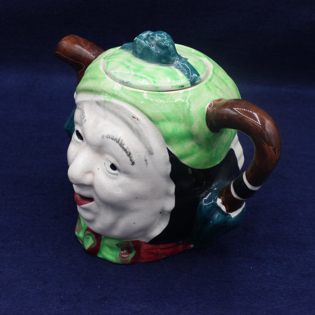 Ceramic teapot shaped like a face with a green lid and brown handle on a blue background