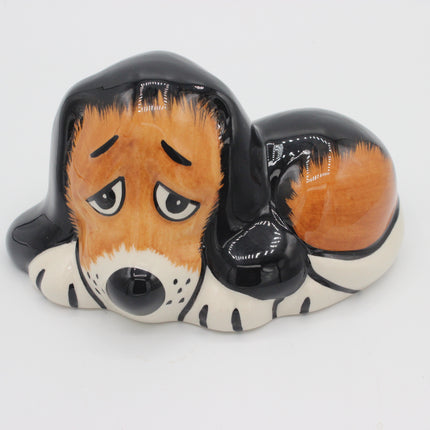 A ceramic dog figurine or piggy bank shaped like a sleeping basset hound, with a glossy black and orange-brown coat, white paws and muzzle, and a sad expression, photographed on a white background.