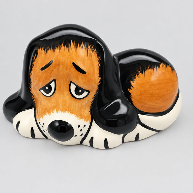 A ceramic dog figurine or piggy bank shaped like a sleeping basset hound, with a glossy black and orange-brown coat, white paws and muzzle, and a sad expression, photographed on a white background.