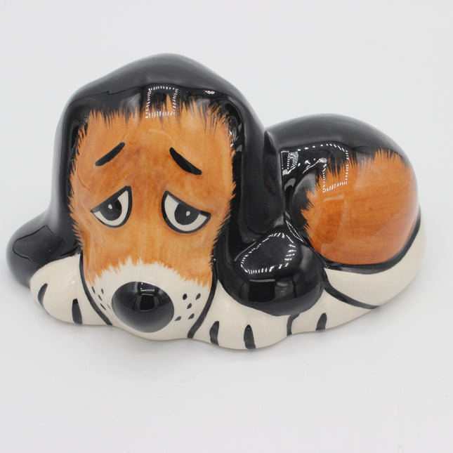 A ceramic dog figurine or piggy bank shaped like a sleeping basset hound, with a glossy black and orange-brown coat, white paws and muzzle, and a sad expression, photographed on a white background.
