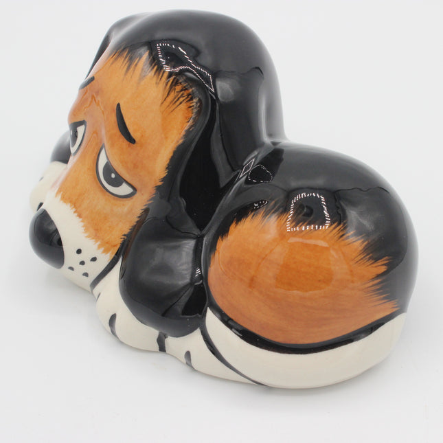 A glossy ceramic dog-shaped container or figurine with a black body, orange and cream fur pattern, and a small window on the back, shown on a plain white background.