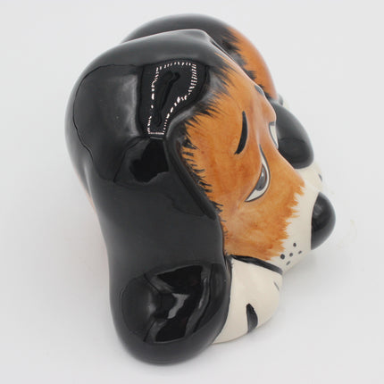 A glossy ceramic dog head figurine or mask, featuring a black body with orange, white, and black painted details on its face, shown against a plain white background.