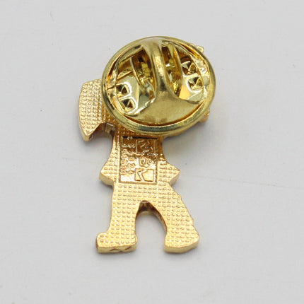 A small gold-tone pin or charm shaped like a stylized figure, shown from the back with its metal clasp visible, on a plain light gray background.