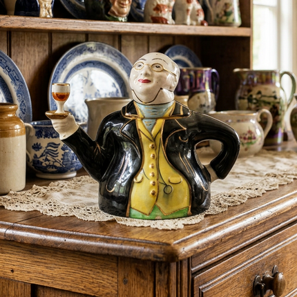 A ceramic teapot-shaped figurine of a man in a black coat with a yellow vest and green trousers, holding a glass of amber liquid, displayed on a wooden cabinet with vintage pottery in the background.